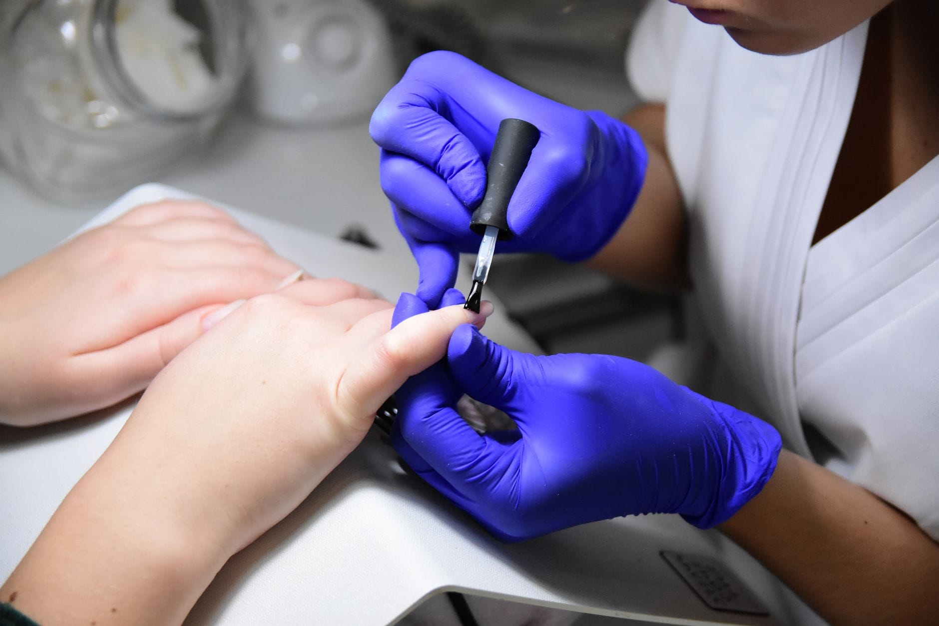 Manicurzystka w fioletowych rękawiczkach nakłada lakier na paznokcie klientki